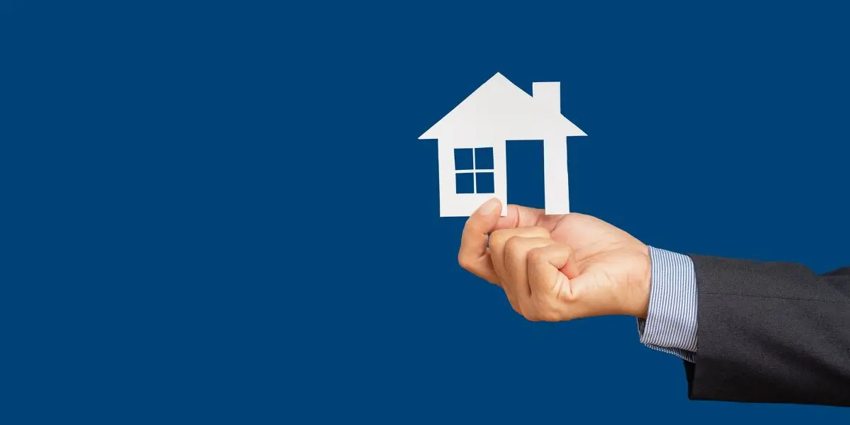 Uma mão segurando o recorte de uma casa em um fundo azul